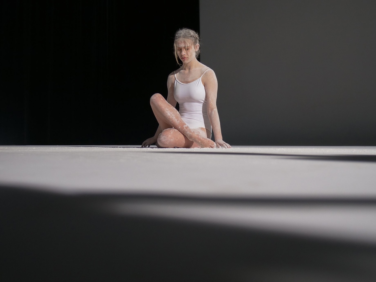 Dancer seated on a white studio floor cut by diagonal shadows
